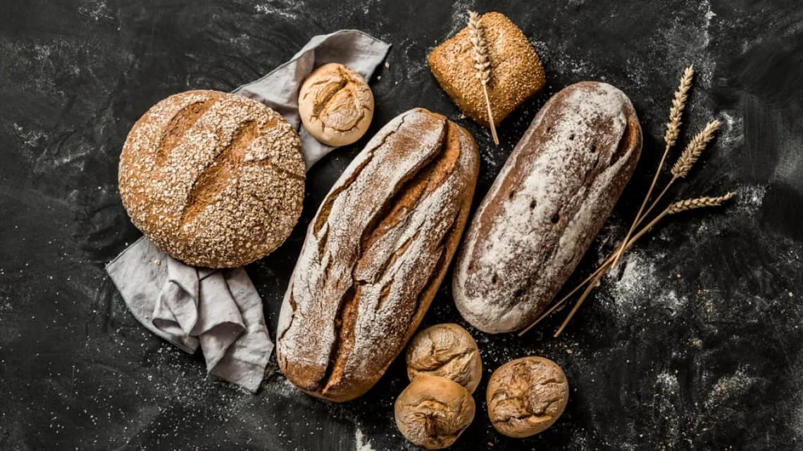 Golden brown loaf of bread on a rustic wooden table, surrounded by wheat stalks and a linen cloth, evoking a warm and inviting bakery atmosphere. The mood is cozy and welcoming. Text on the image: DELIBAKE.